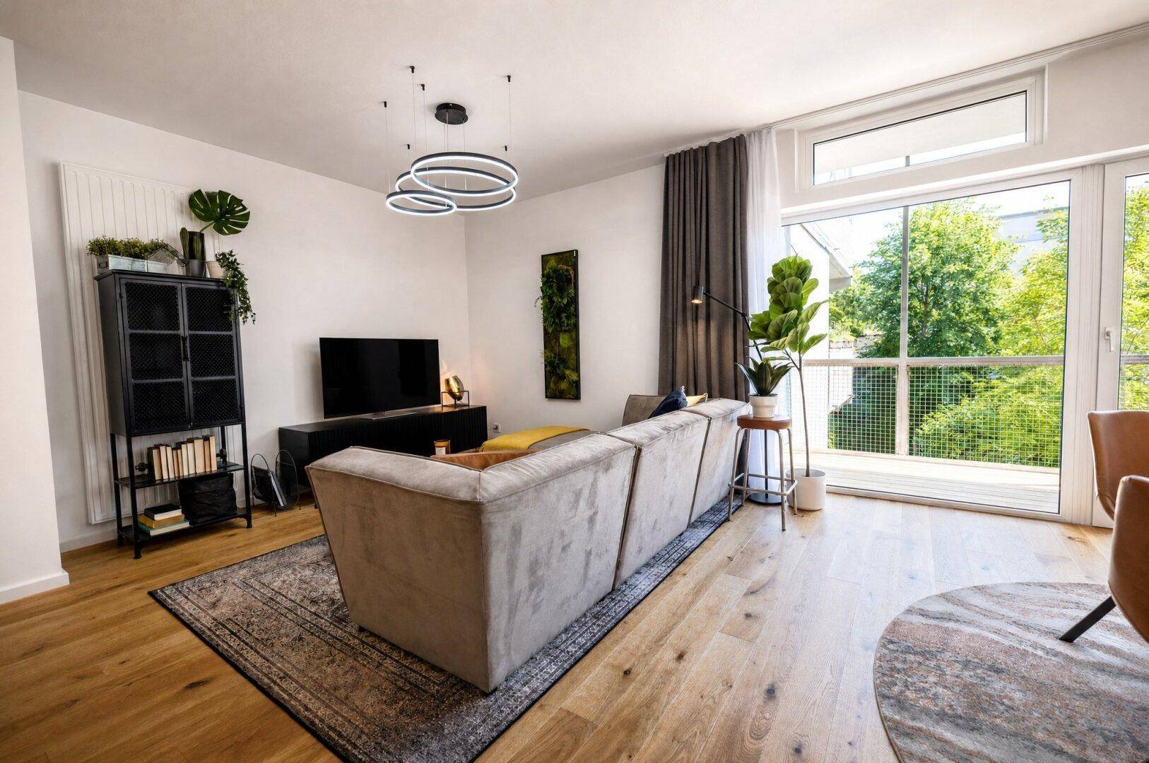 Heller Wohnbereich mit beigem Sofa, Fernseher und weitem Blick ins Grüne.