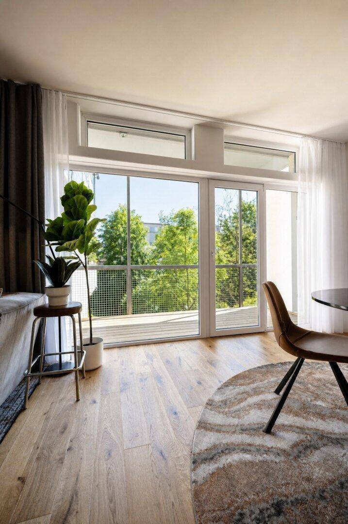 Große Fensterfront mit Zugang zum Balkon und Blick auf grüne Bäume.