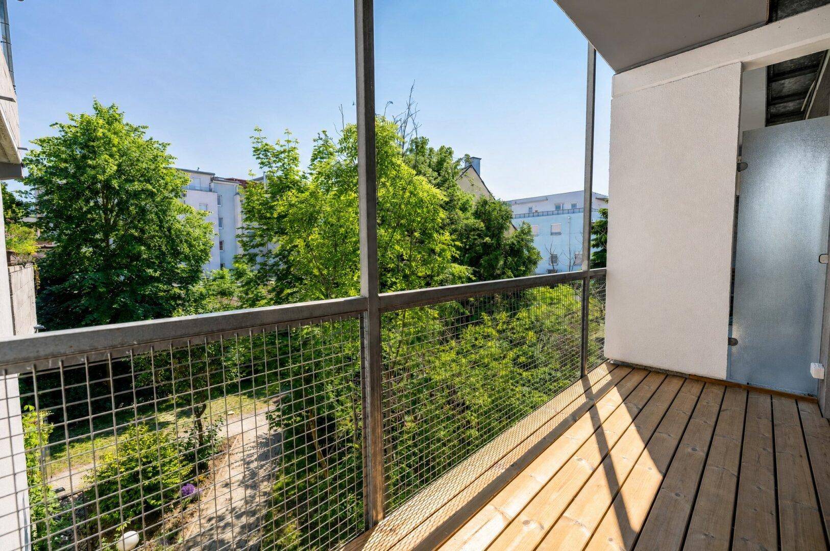 Sonniger Balkon mit Holzboden und Blick auf Bäume und umliegende Gebäude.