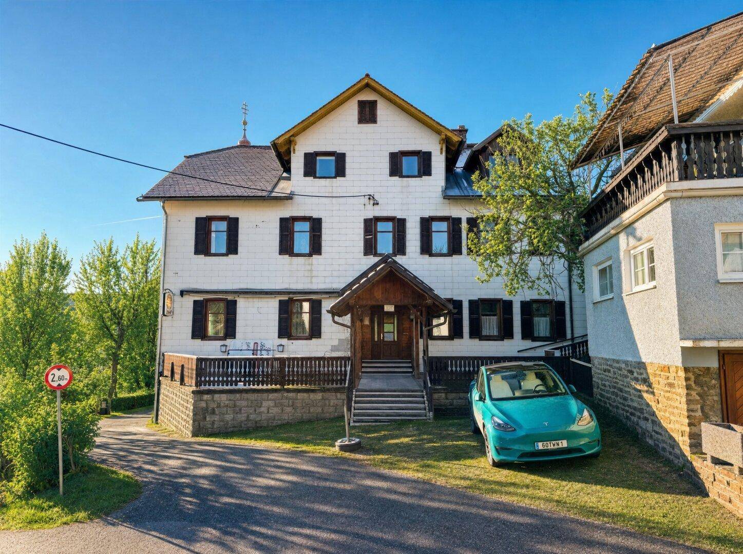 Historisches Gebäude mit weißer Fassade, Holzfensterläden und einem türkisfarbenen Elektroauto davor.