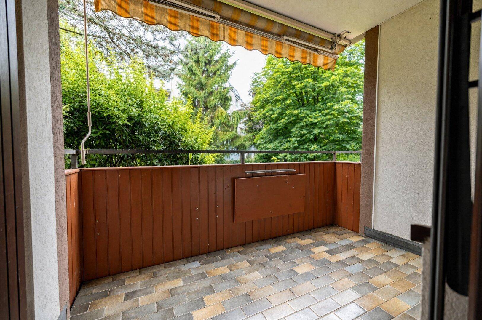 Balkon mit Fliesenboden und Markise, umgeben von viel Grün.