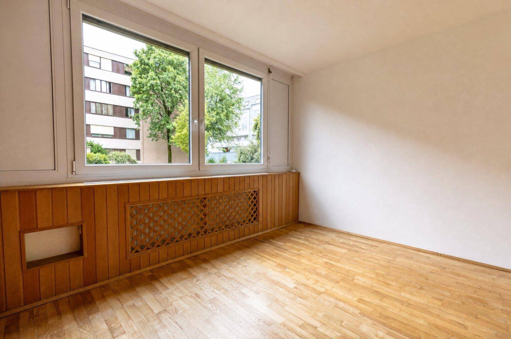 Heller Raum mit Parkettboden und großen Fenstern mit Holzverkleidung.