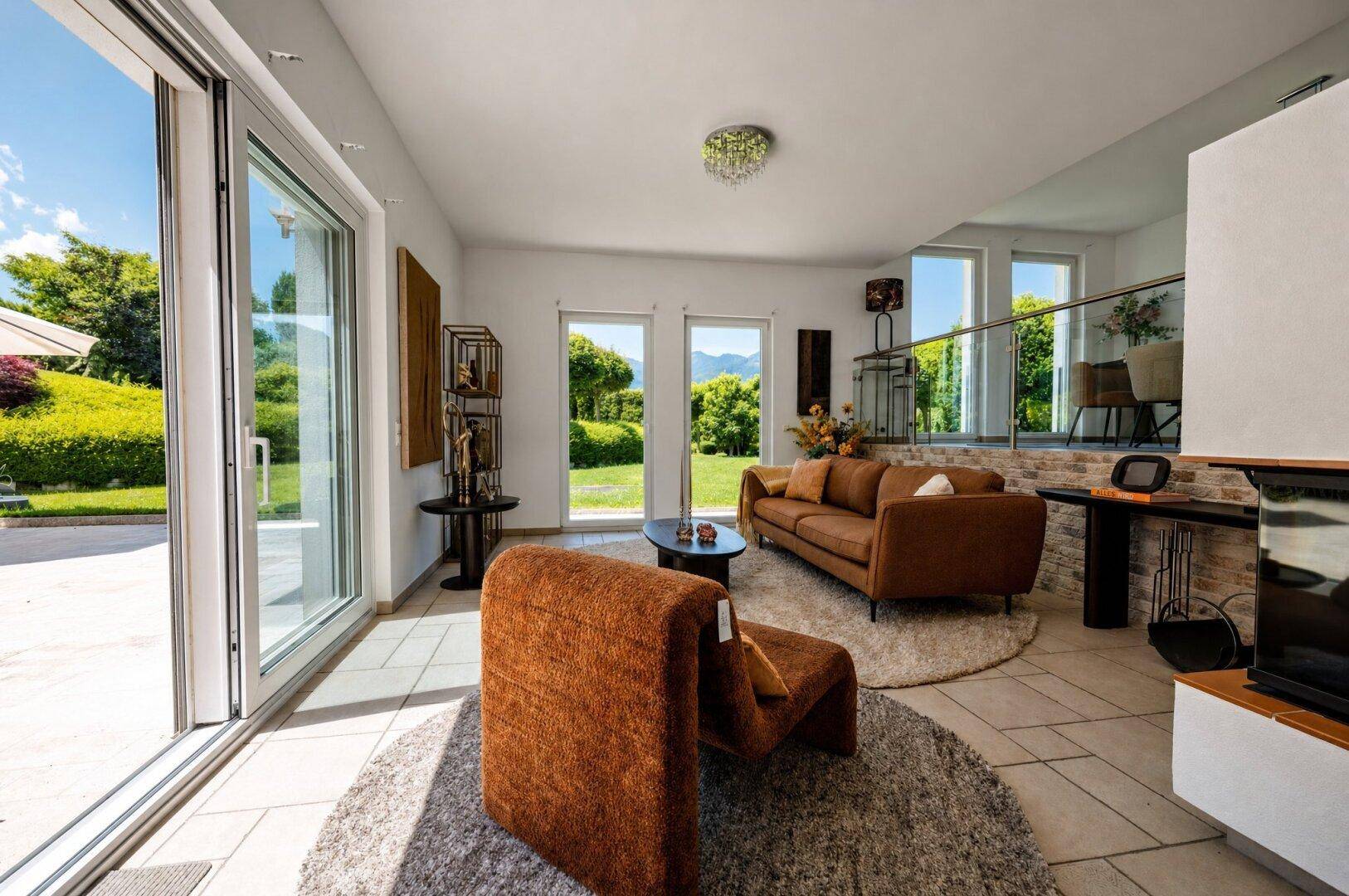 Gemütliches Wohnzimmer mit braunem Sofa, Teppich und großen Fenstern mit Zugang zum Garten.