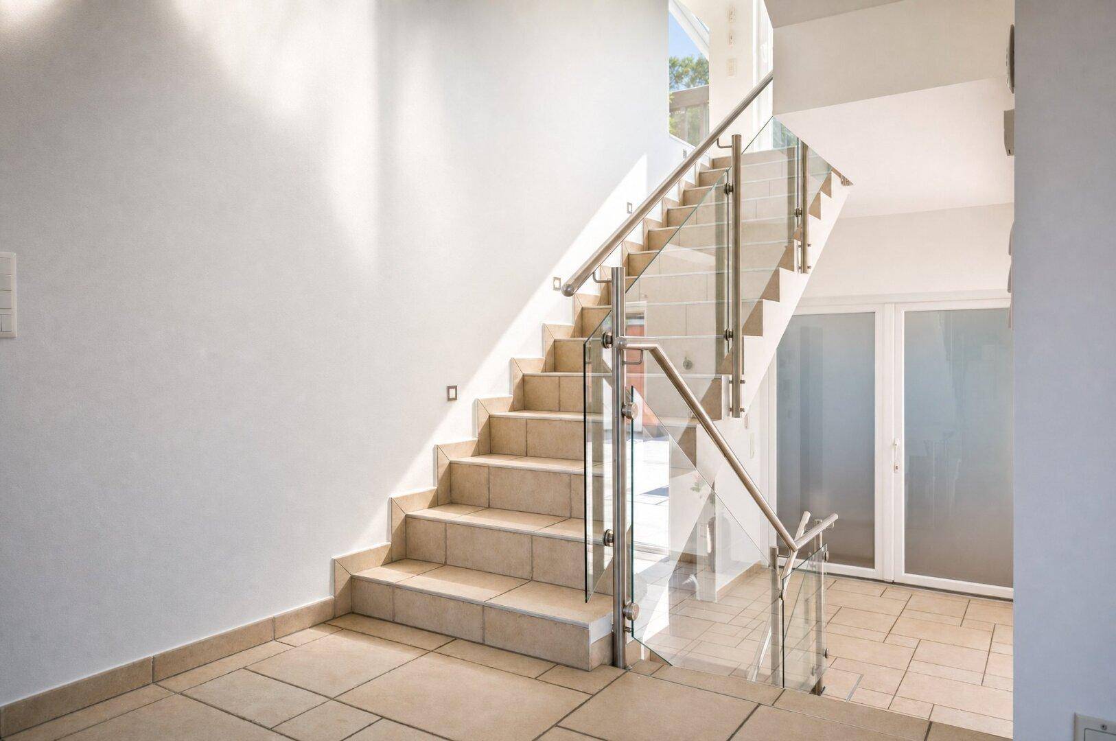 Moderne Treppe mit Glasgeländer und Edelstahlhandlauf in einem hellen Flurbereich.