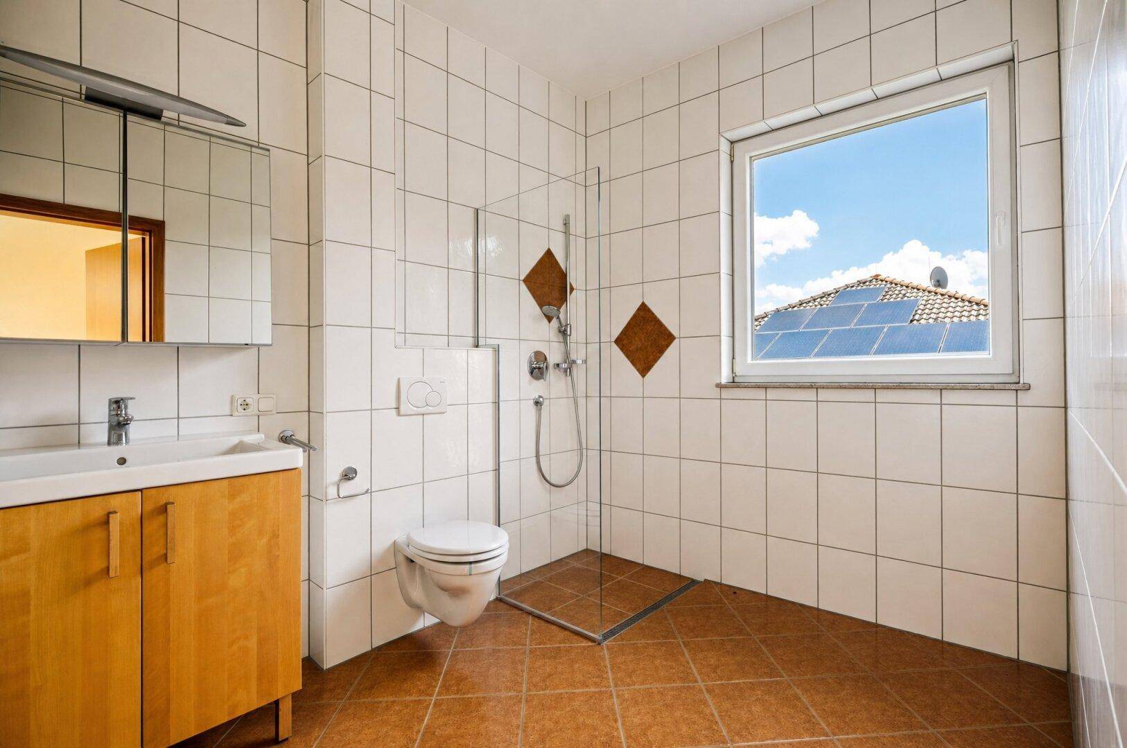 Helles Badezimmer mit weißen Fliesen, ebenerdiger Dusche und Fenster mit Blick auf Solarpaneele.