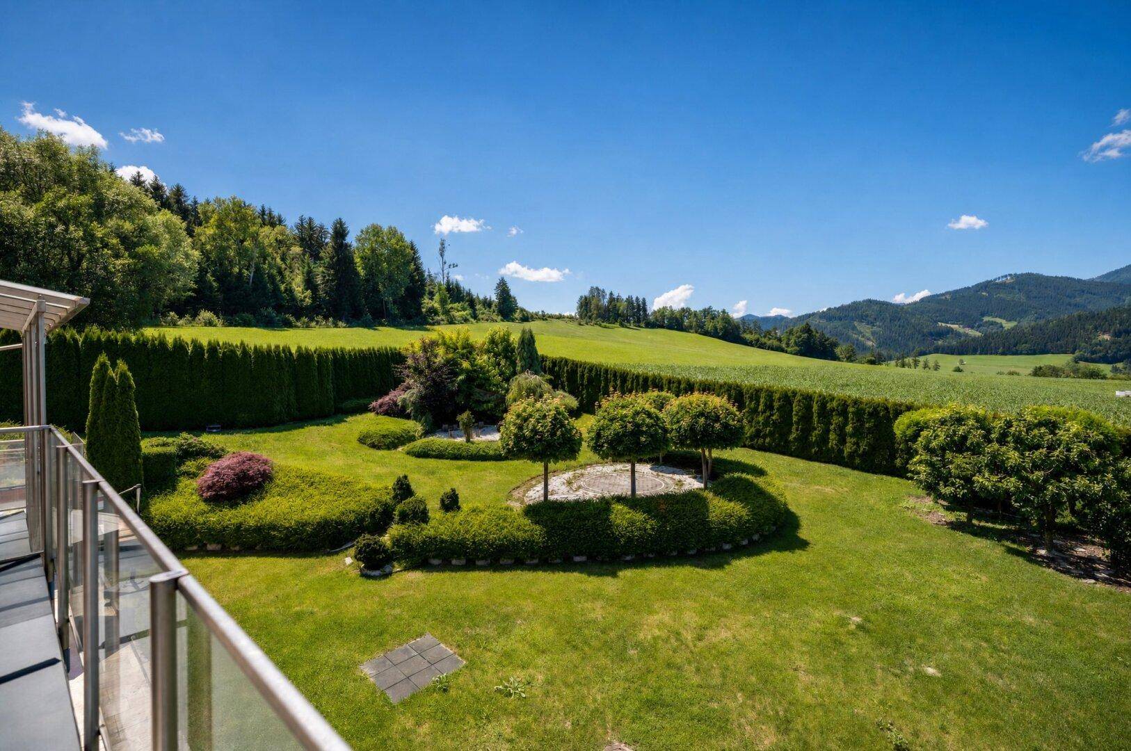 Gepflegter Garten mit Rasenflächen, hohen Hecken und dekorativen Bäumen, umgeben von grünen Feldern.