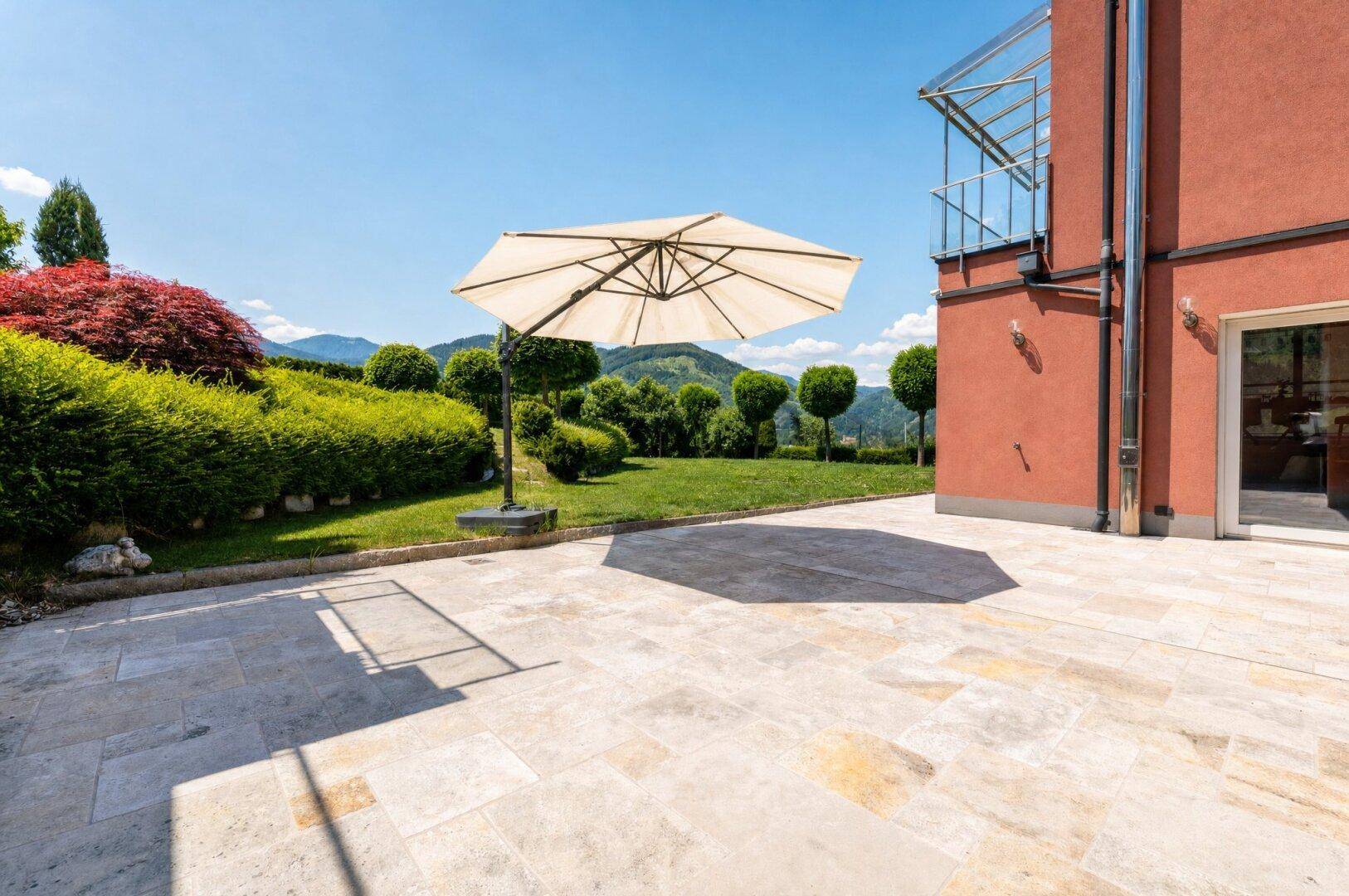 Großzügige gepflasterte Terrasse mit Sonnenschirm und Blick auf den gepflegten Garten und die Berge.