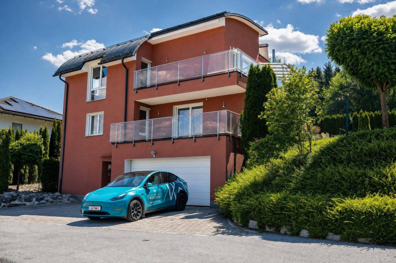 Modernes Wohnhaus mit mehreren Balkonen, integrierter Garage und einem Elektroauto in der Einfahrt.