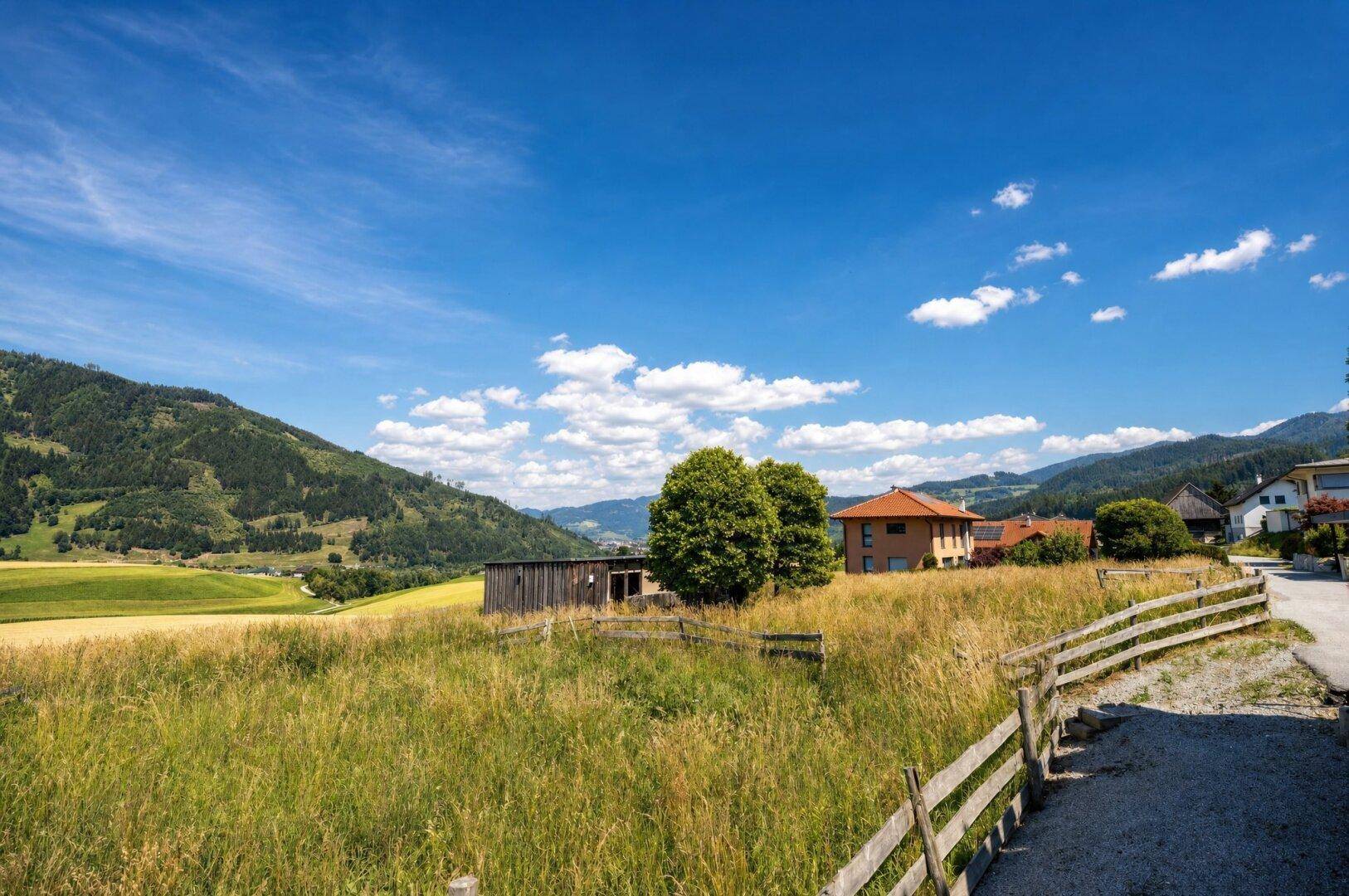 Weitläufige Landschaft mit grünen Feldern, bewaldeten Bergen und vereinzelten Häusern in der Ferne.