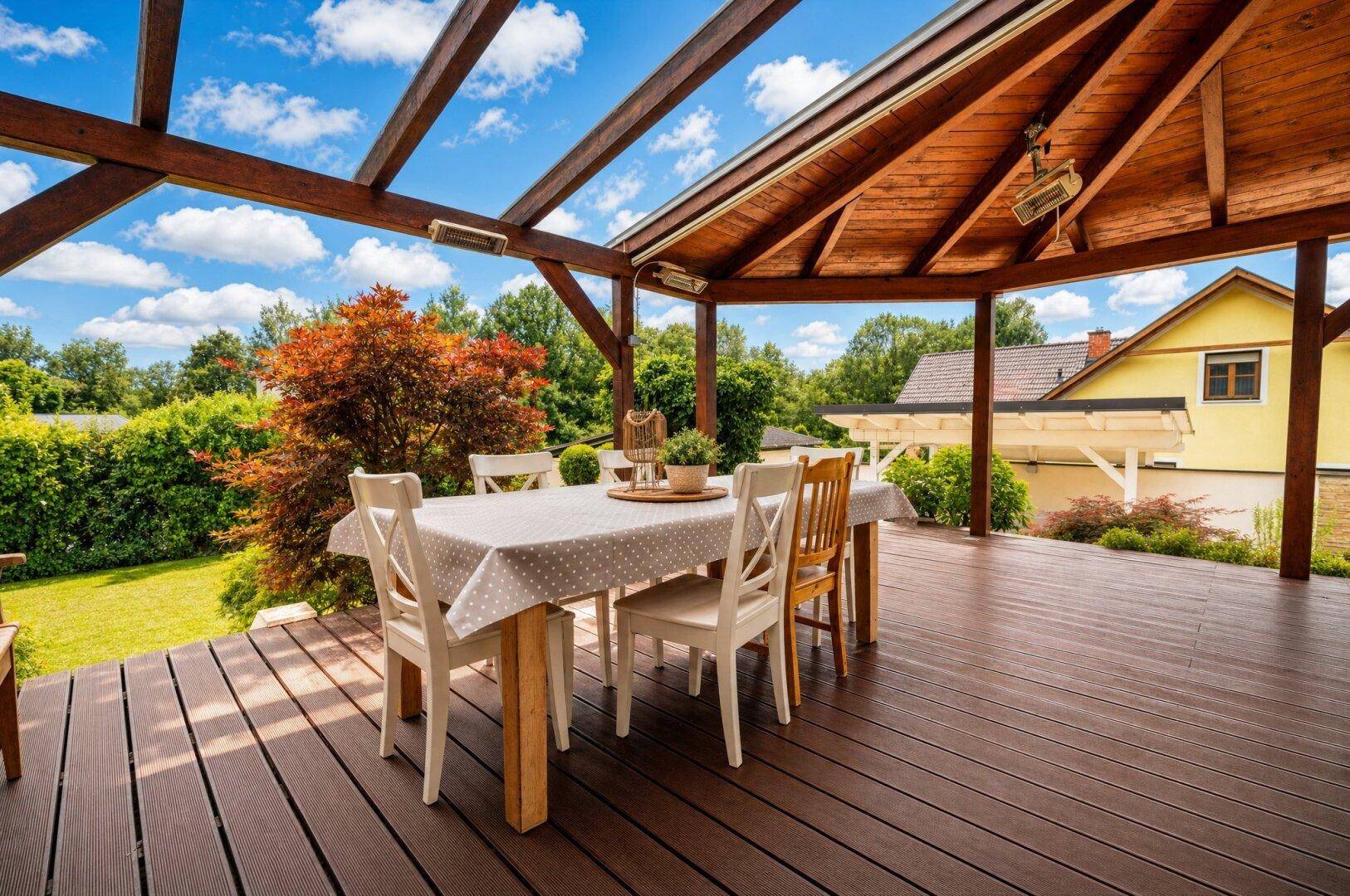 Überdachte Holzterrasse mit Esstisch und Stühlen, umgeben von üppigem Grün und Gartenblick.