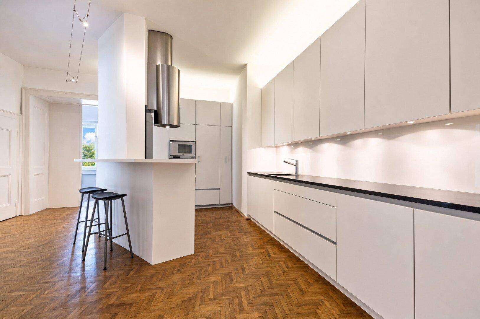 Moderne Einbauküche mit weißen Schränken, dunkler Arbeitsplatte und Kochinsel mit Barhockern.
