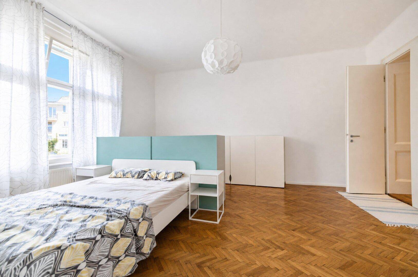 Helles Schlafzimmer mit Fischgrätparkett, großem Fenster und minimalistischer Einrichtung.