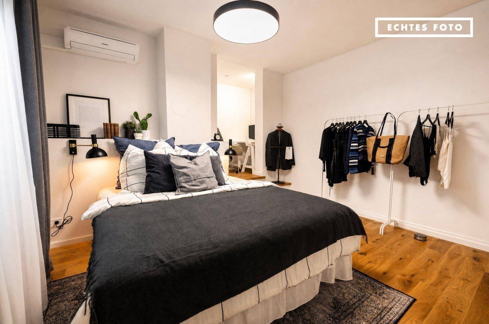 Helles Schlafzimmer mit Holzboden, Klimaanlage und offenem Kleiderständer für Kleidung und Accessoires.