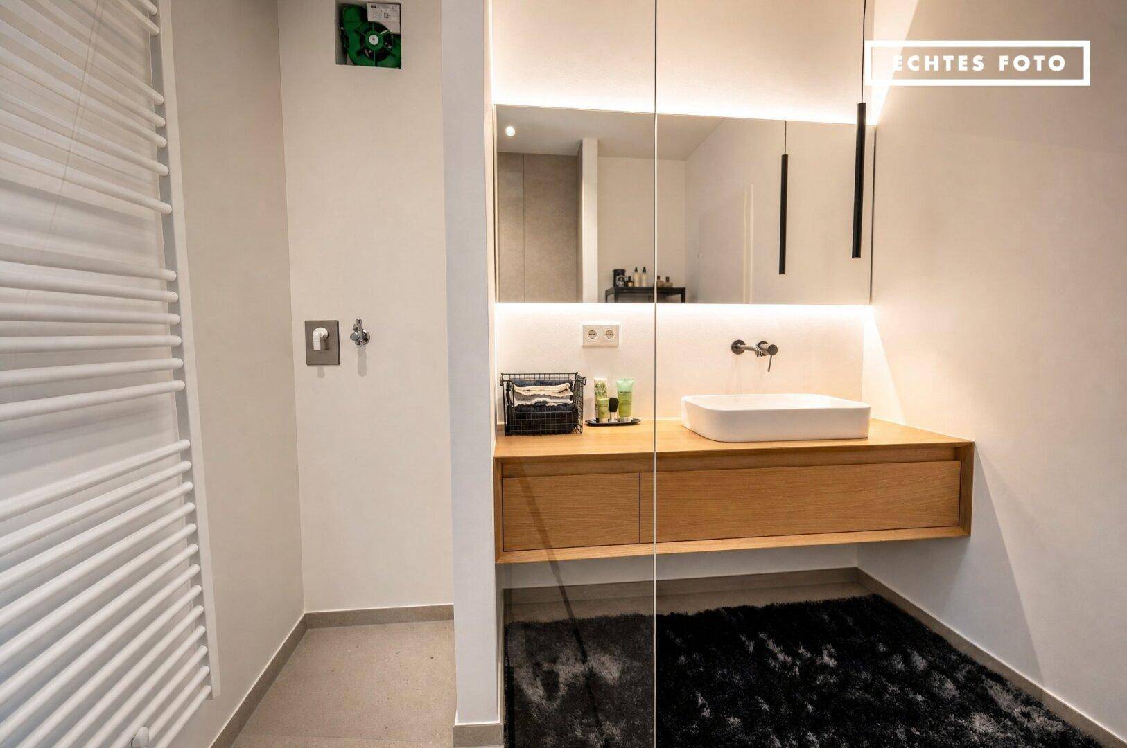Modernes Badezimmer mit Holzwaschtisch, beleuchtetem Spiegel und weißem Aufsatzwaschbecken.