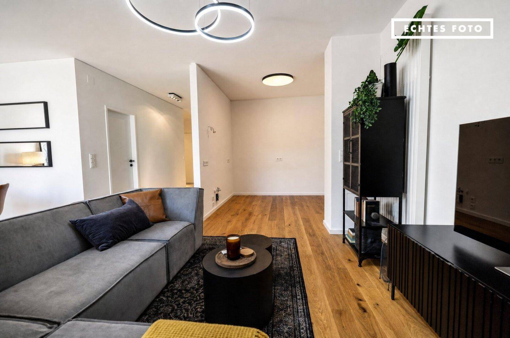 Moderner Wohnbereich mit grauem Sofa, Holzboden und Übergang zu einem leeren, hellen Raum.