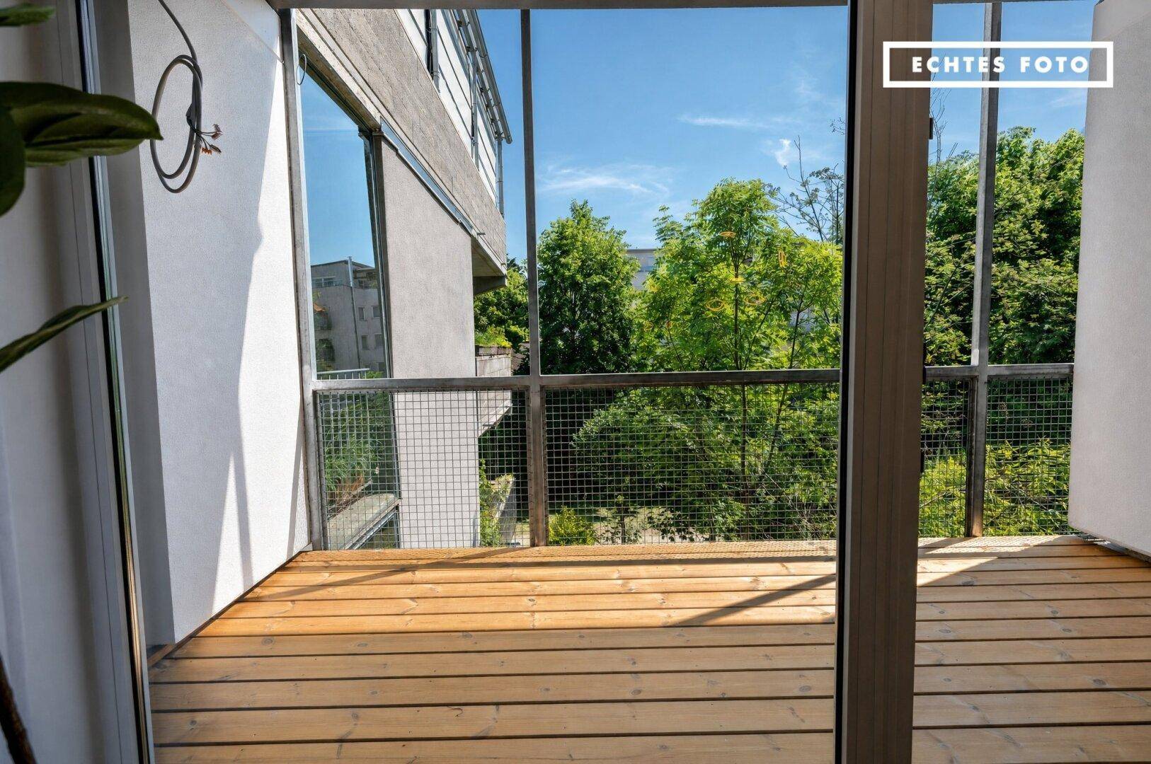 Heller Balkon mit Holzboden und Gitterbrüstung, umgeben von Bäumen und mit Blick ins Grüne.