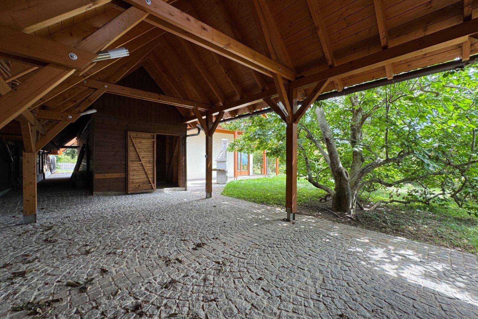 Geräumiger Carport mit Kopfsteinpflaster und Holzbalkenkonstruktion, angrenzend an einen Baum.