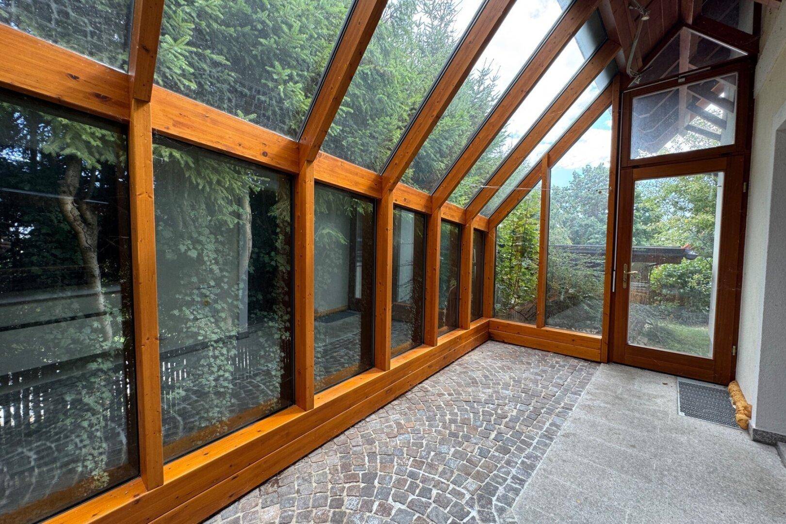 Heller Wintergarten mit Glasdach und Steinboden, umgeben von viel Grün.