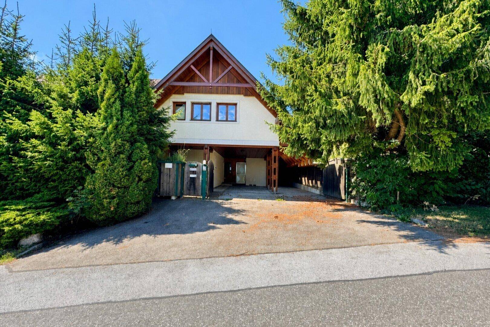 Einfamilienhaus mit Carport und gepflegtem Vorgarten, umgeben von hohen Bäumen.