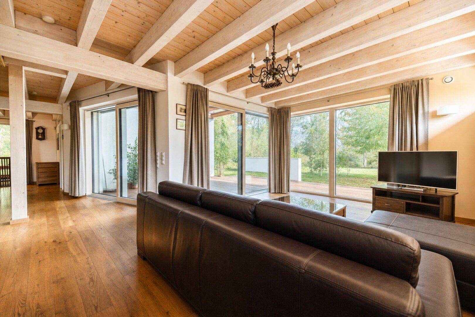 Geräumiges Wohnzimmer mit großen Fensterfronten, Holzbalkendecke und Zugang zur Terrasse.