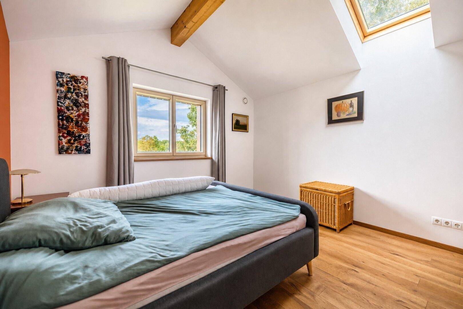 Helles Schlafzimmer mit Doppelbett, Holzbalken, Dachfenster und Blick ins Grüne.