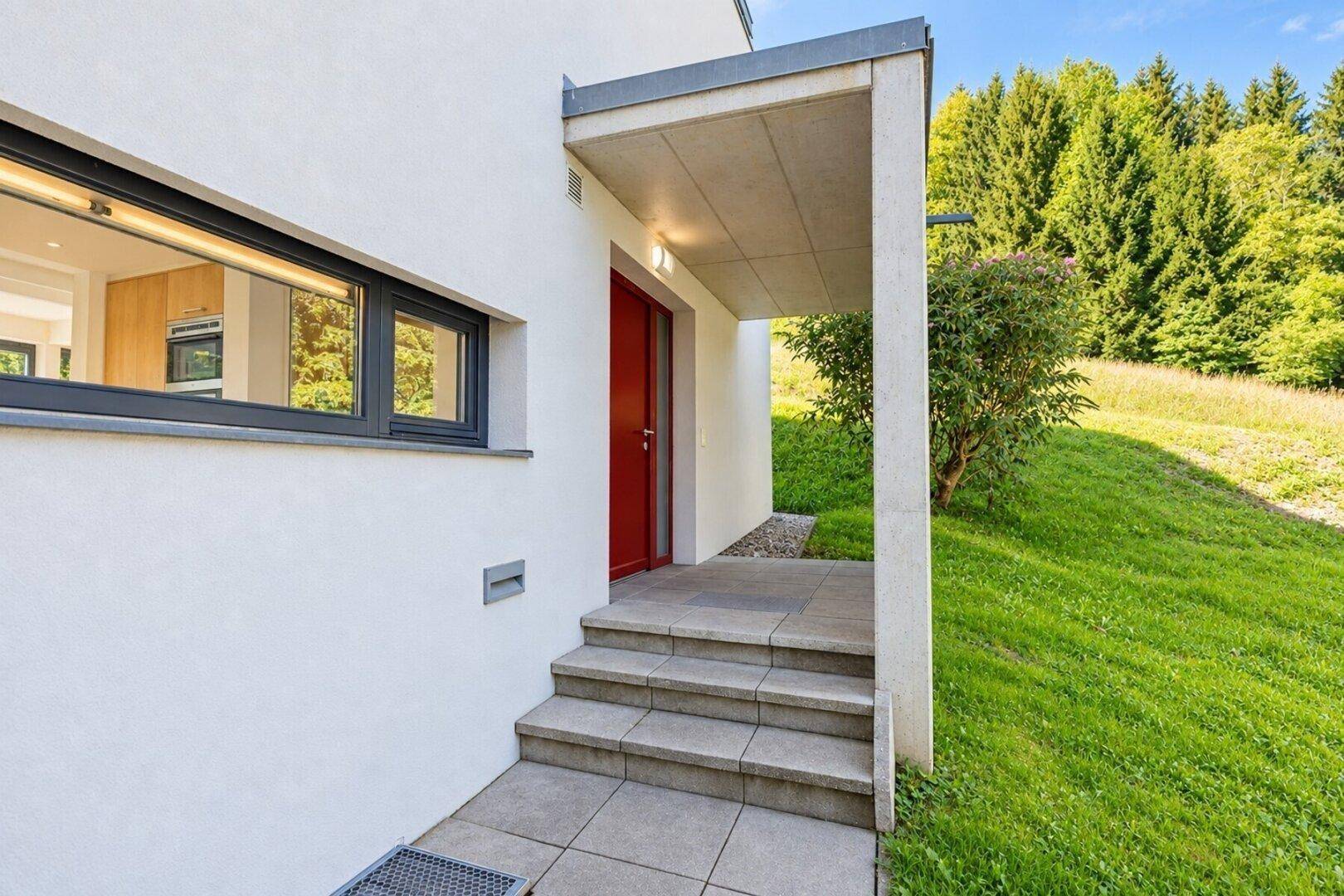 Eingangsbereich des Hauses mit roten Türen und überdachtem Zugang, umgeben von Grünflächen.