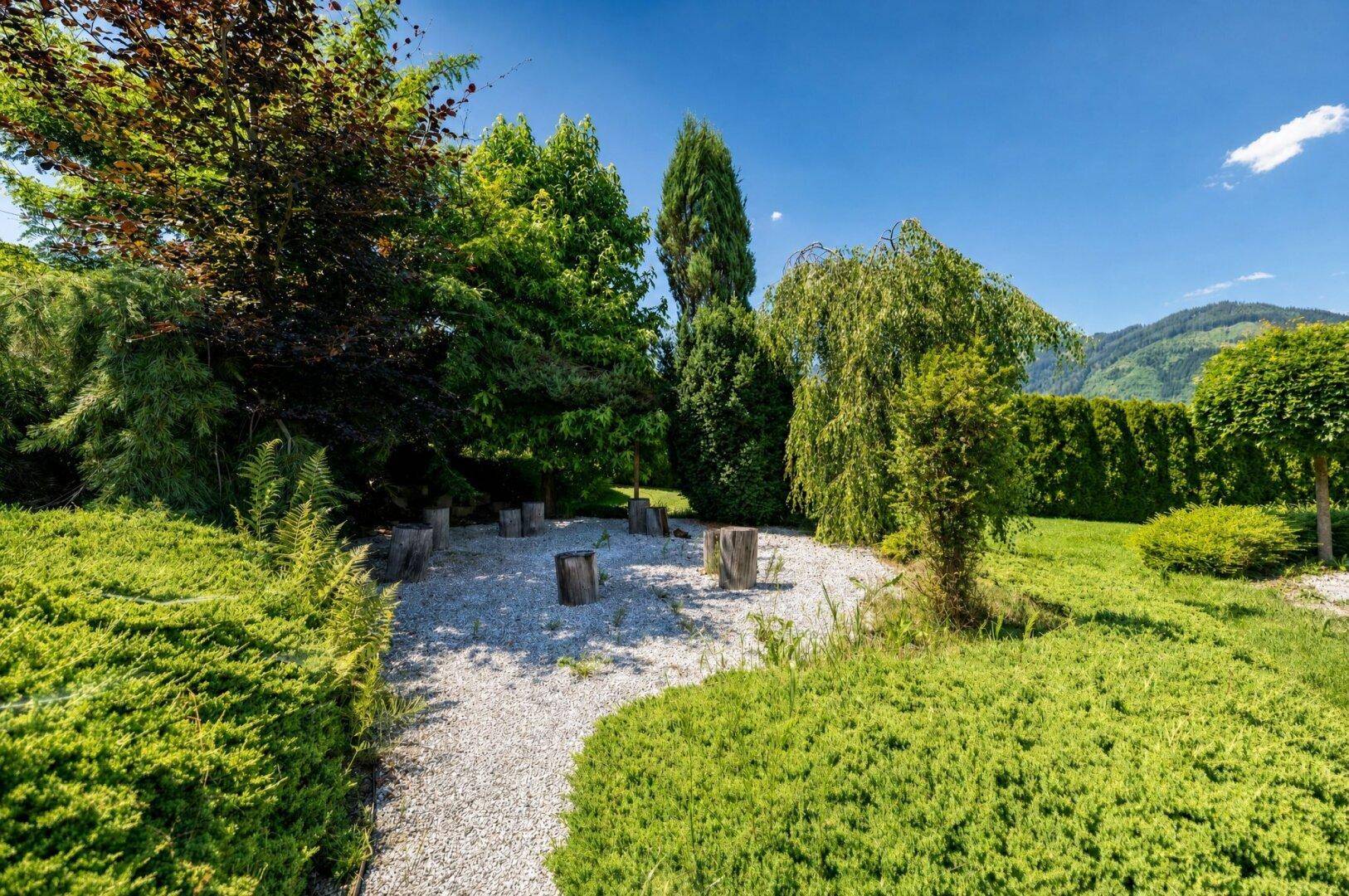 Idyllischer Gartenbereich mit Kiesweg, verschiedenen Bäumen und Baumstümpfen als natürliche Sitzgelegenheiten.
