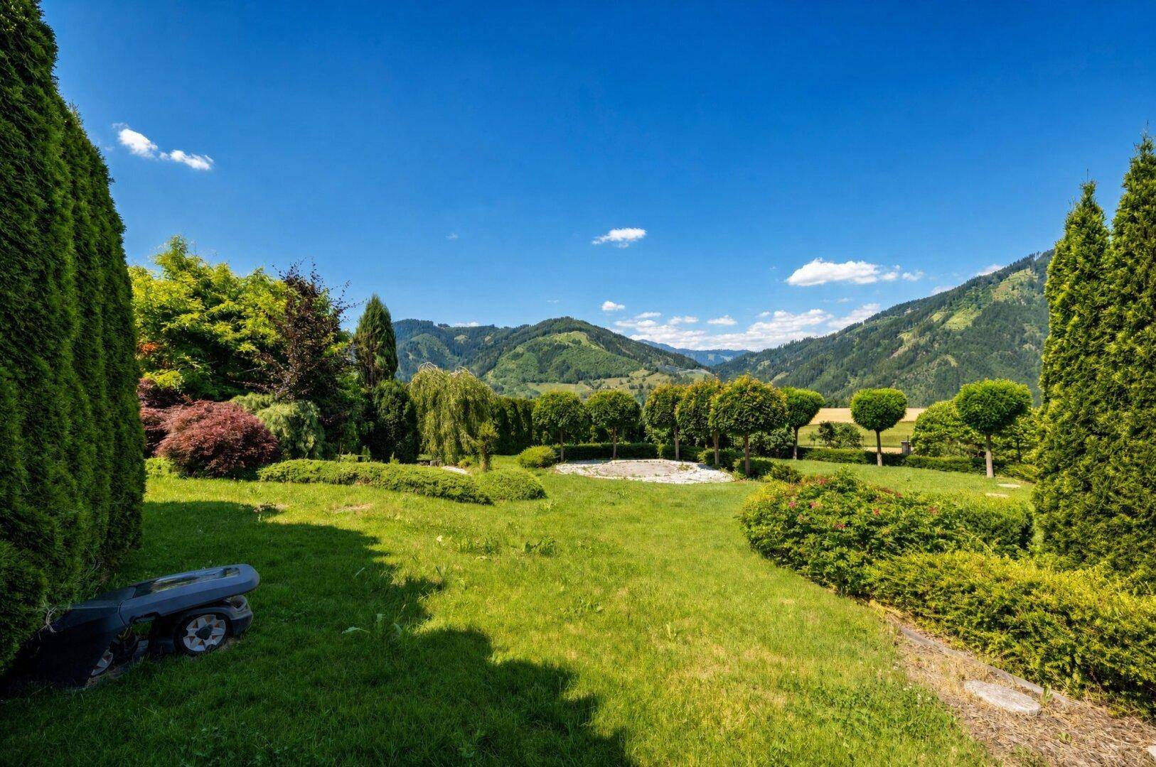 Grüner Garten mit verschiedenen Bäumen und Sträuchern, der einen weiten Blick auf die umliegenden Berge bietet.