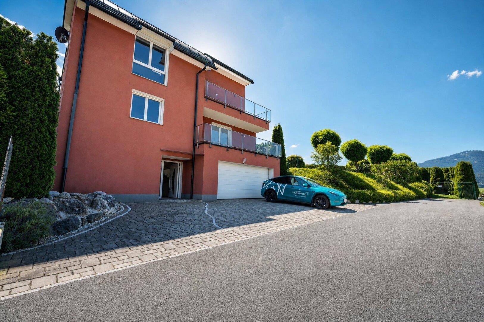 Modernes Wohnhaus mit roter Fassade, integrierter Garage und gepflasterter Auffahrt unter blauem Himmel.