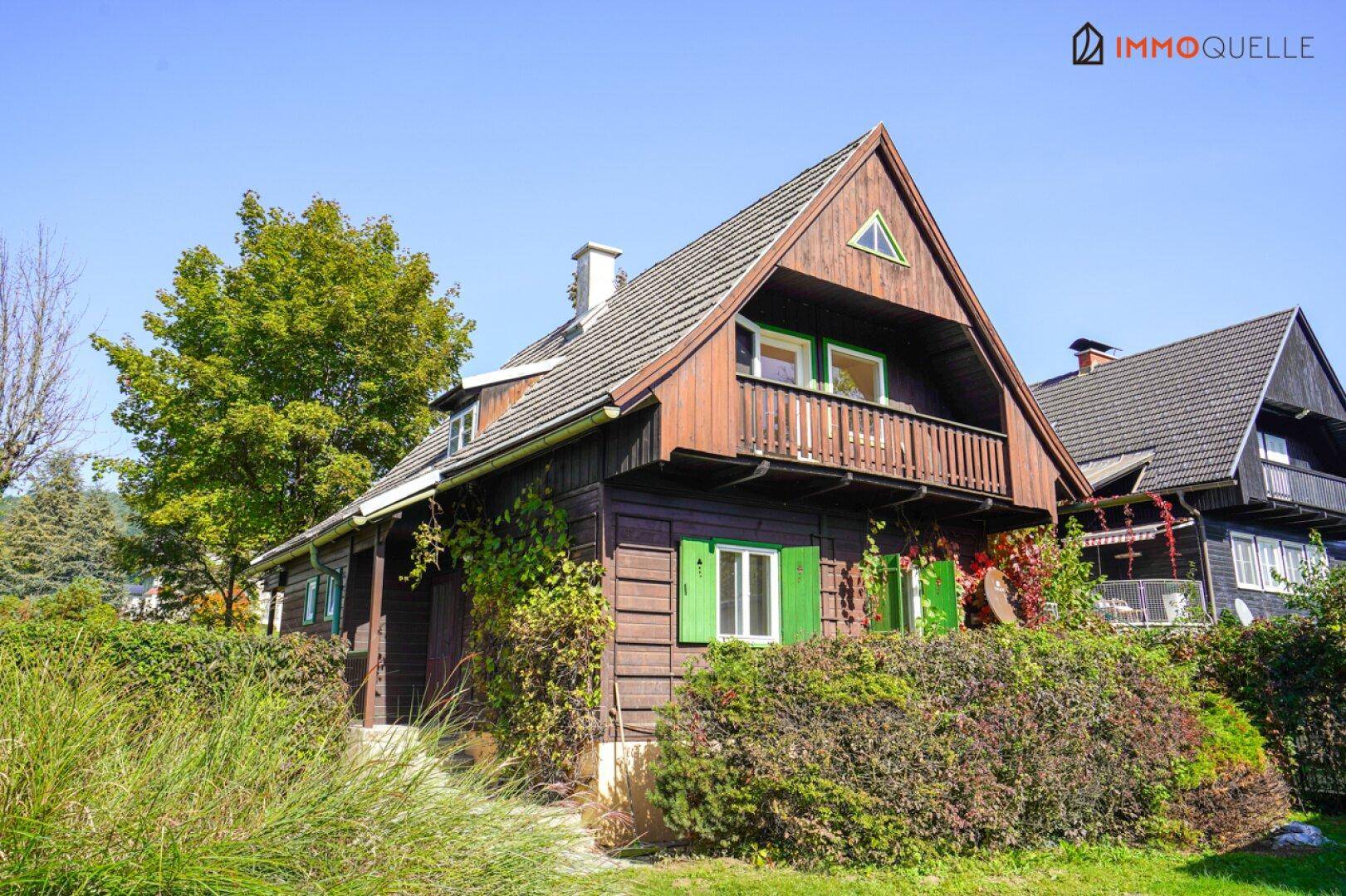 Traditionelles Holzhaus mit Balkon, grünen Fensterläden und üppiger Vegetation im Garten.