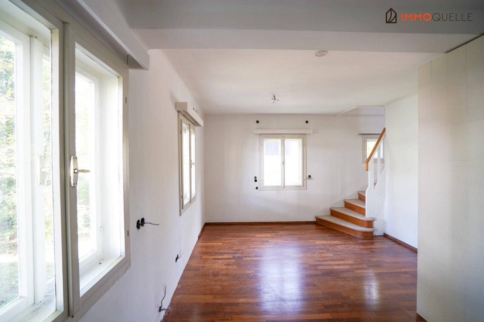 Leerer Raum mit Holzboden, großen Fenstern und Treppe, bereit für die individuelle Gestaltung.