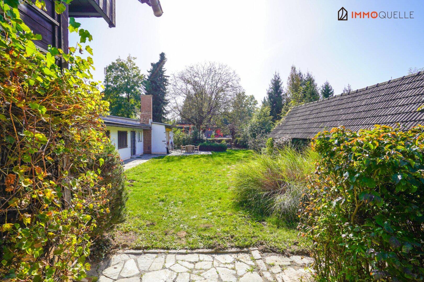 Weitläufiger Gartenbereich mit Rasen, Sträuchern und Bäumen, umgeben von Gebäuden unter blauem Himmel.