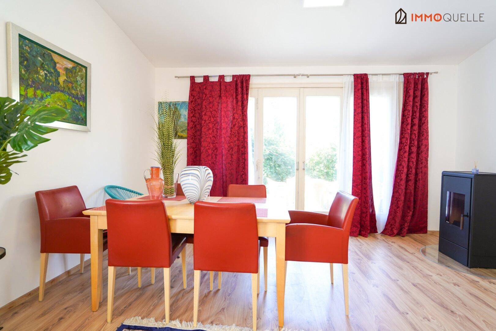 Essbereich mit Holztisch, roten Stühlen und Laminatboden, hell beleuchtet durch große Fenster.