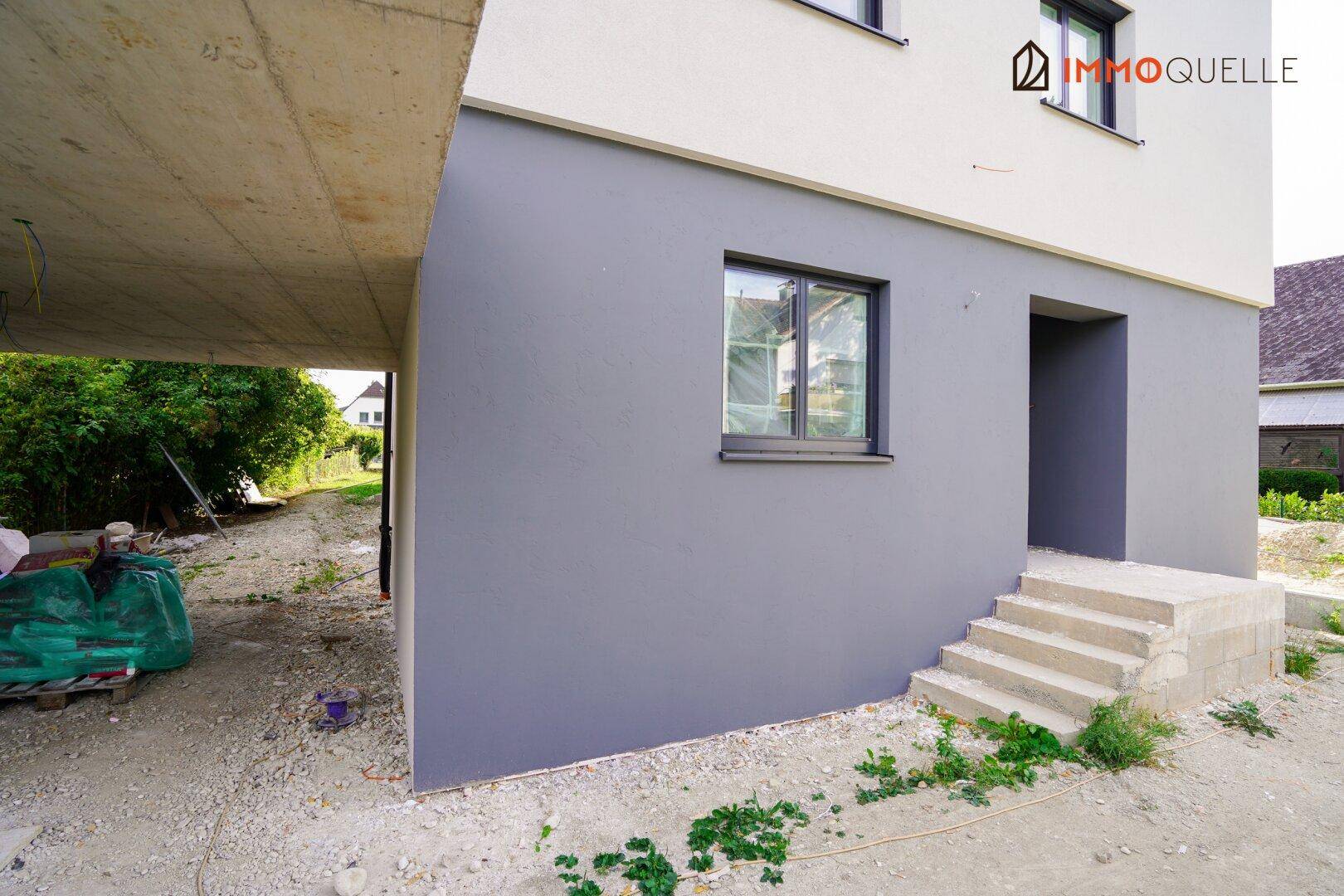 Unfertige Hausfassade mit Eingangsbereich und Fenster, umgeben von Baustellengelände.