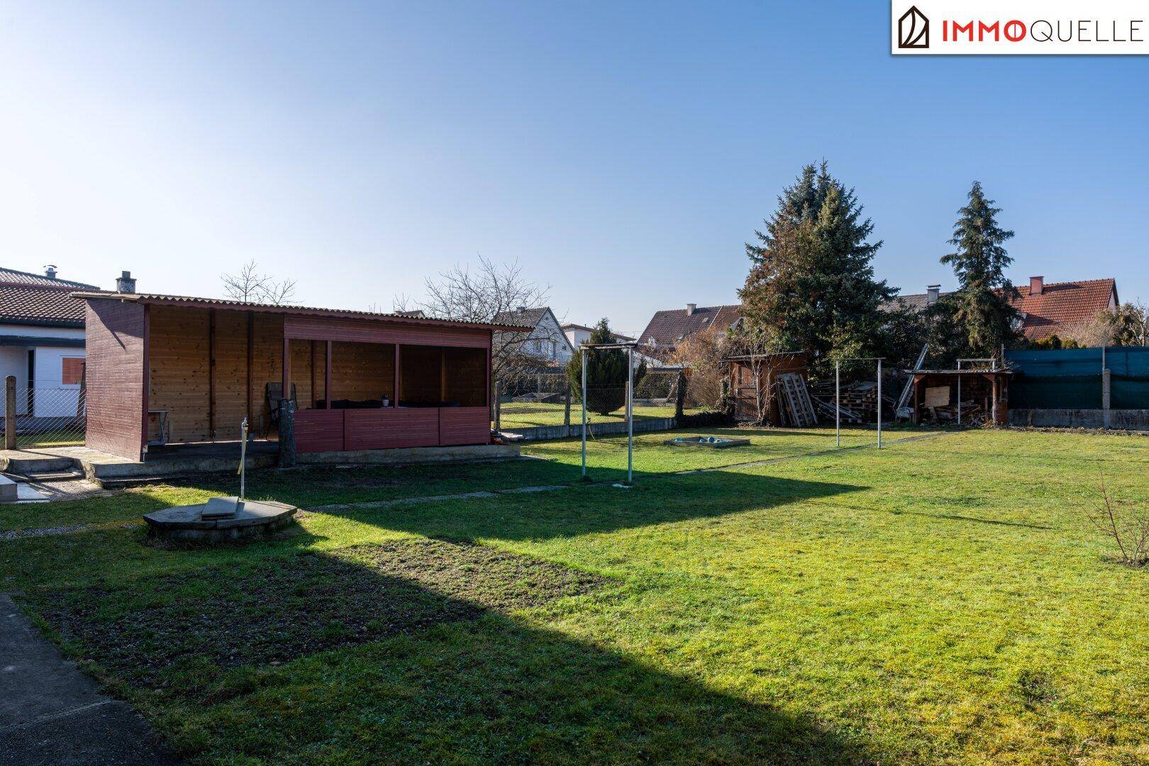 Großer, sonniger Garten mit Rasenfläche und einem überdachten Sitzbereich im Freien.