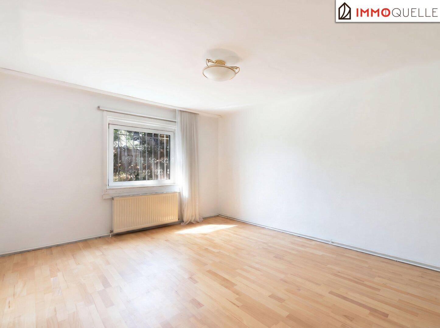 Leeres Wohnzimmer mit hellem Parkettboden und einem großen Fenster mit Blick ins Grüne.