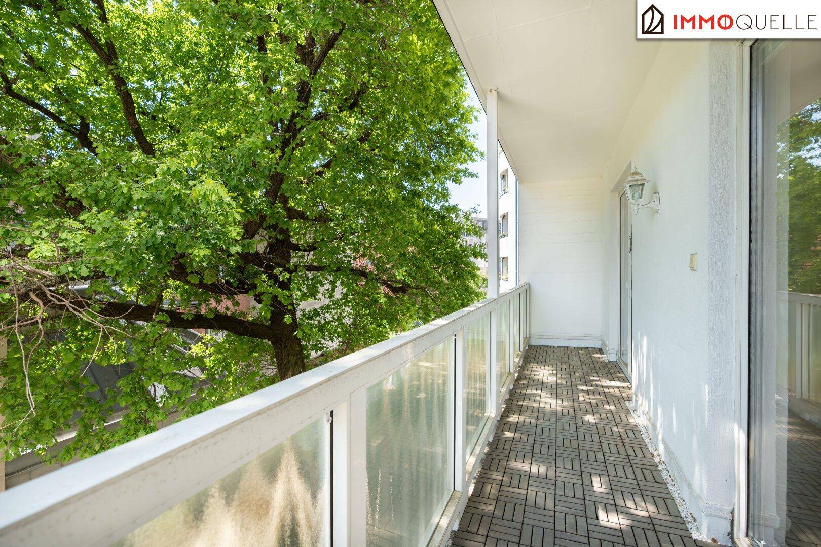 Heller Balkon mit Holzbodenfliesen und üppigem Baumbestand, der Schatten spendet.