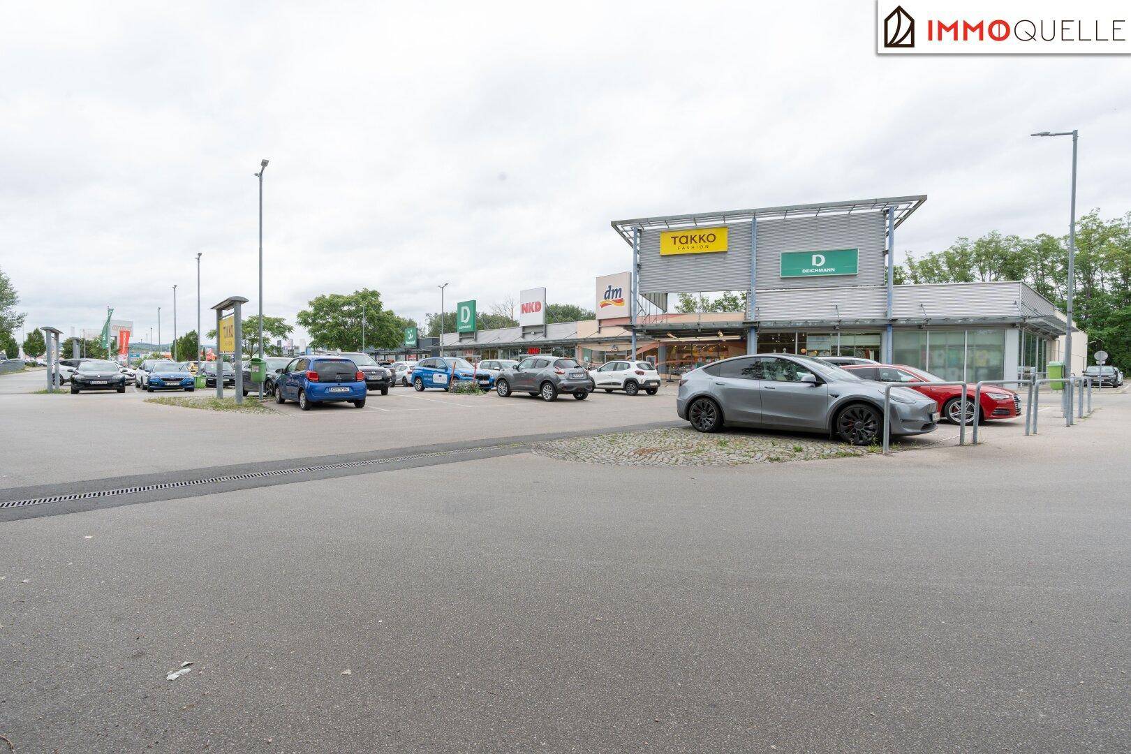 Weitläufiger Parkplatz eines Einkaufszentrums mit verschiedenen Geschäften und vielen geparkten Autos.