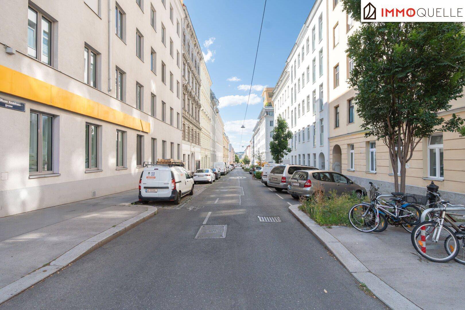 Lange, gerade Straße mit Gebäuden auf beiden Seiten, parkenden Lieferwagen und Fahrrädern.