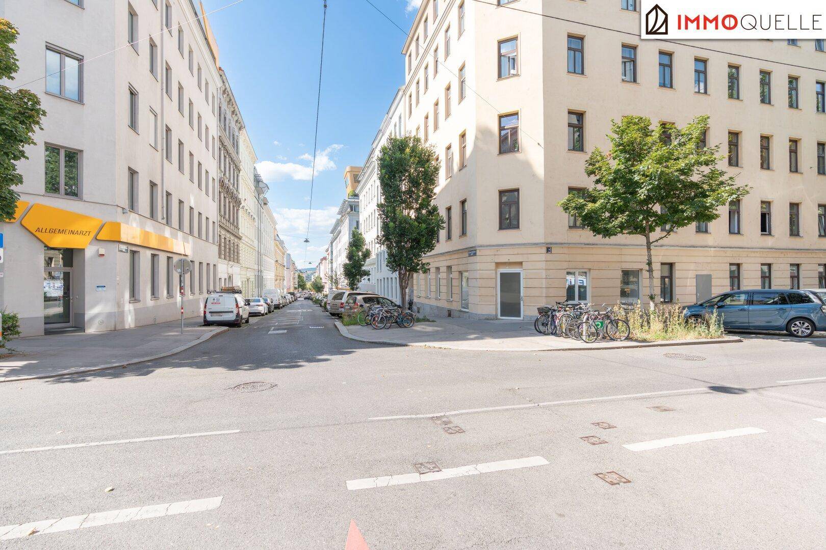 Weitläufige Straßenansicht mit Gebäuden, parkenden Autos und Bäumen unter blauem Himmel.