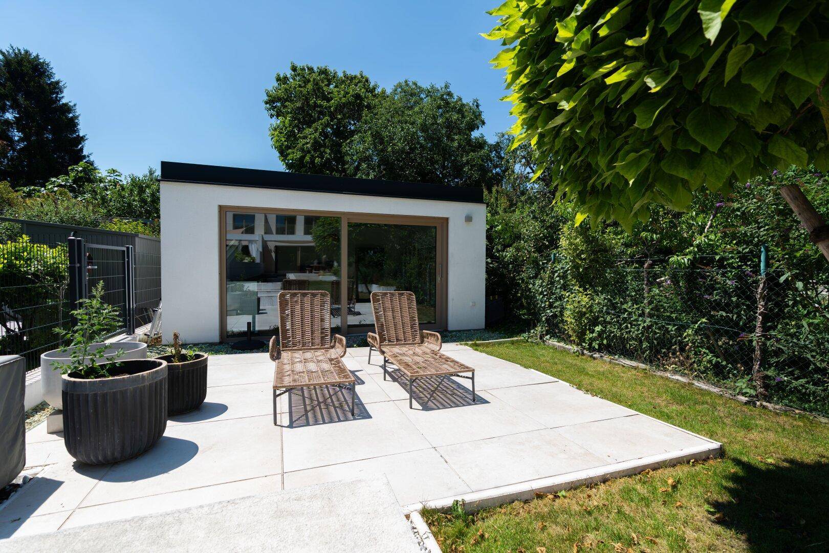 Zwei Liegestühle auf einer gepflasterten Terrasse vor einem modernen Gartenhaus im Grünen.