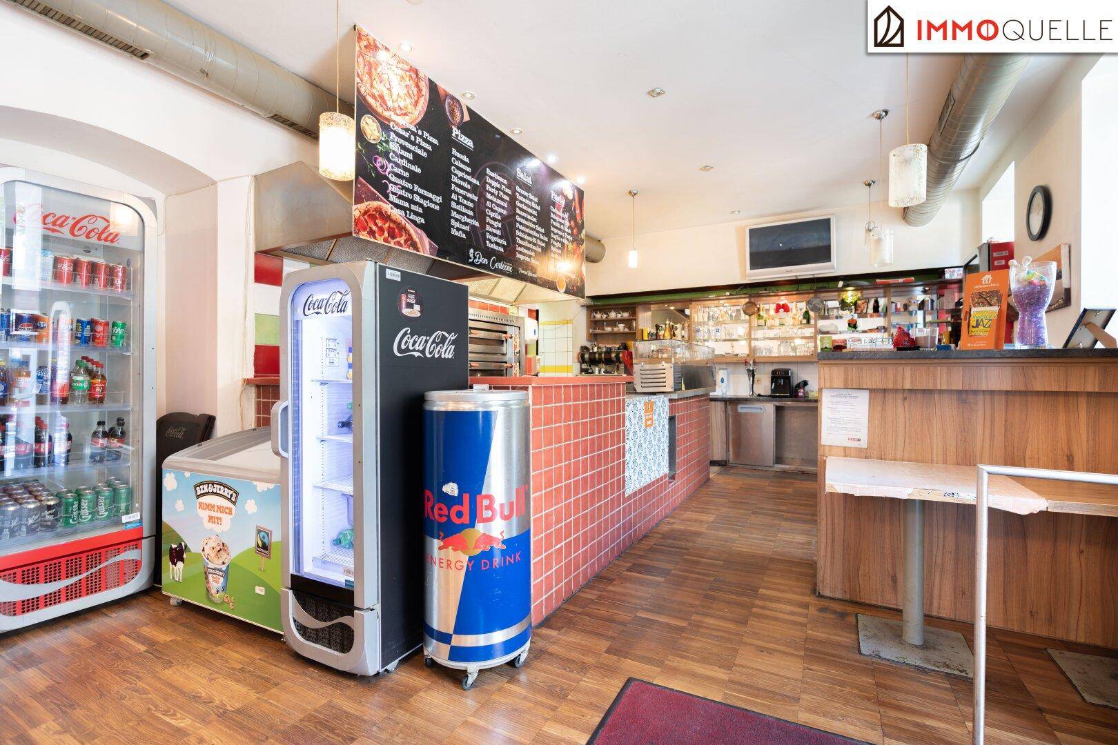 Innenansicht einer Pizzeria mit Theke, offener Küche und mehreren Getränkekühlern.