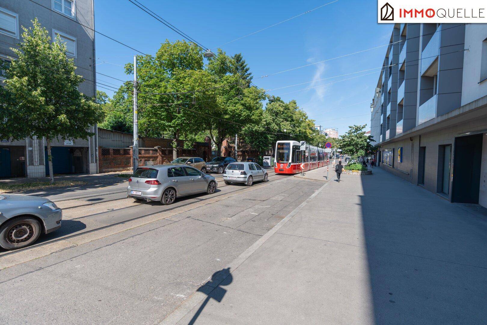 Eine belebte Stadtstraße mit einer roten Straßenbahn, fahrenden Autos und Gebäuden unter blauem Himmel.