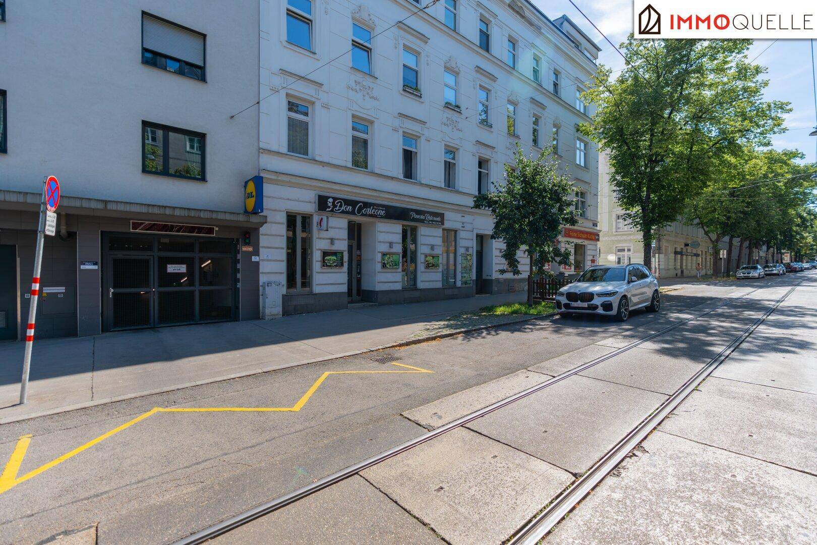 Weitläufige Straßenansicht mit Straßenbahnschienen, geparkten Autos und dem Geschäftsgebäude mit Pizzeria.