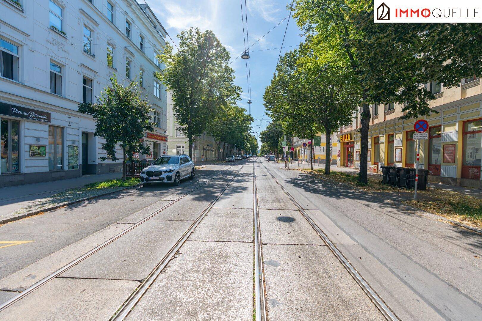 Eine belebte Straße mit Straßenbahnschienen, gesäumt von Bäumen und Wohngebäuden unter blauem Himmel.