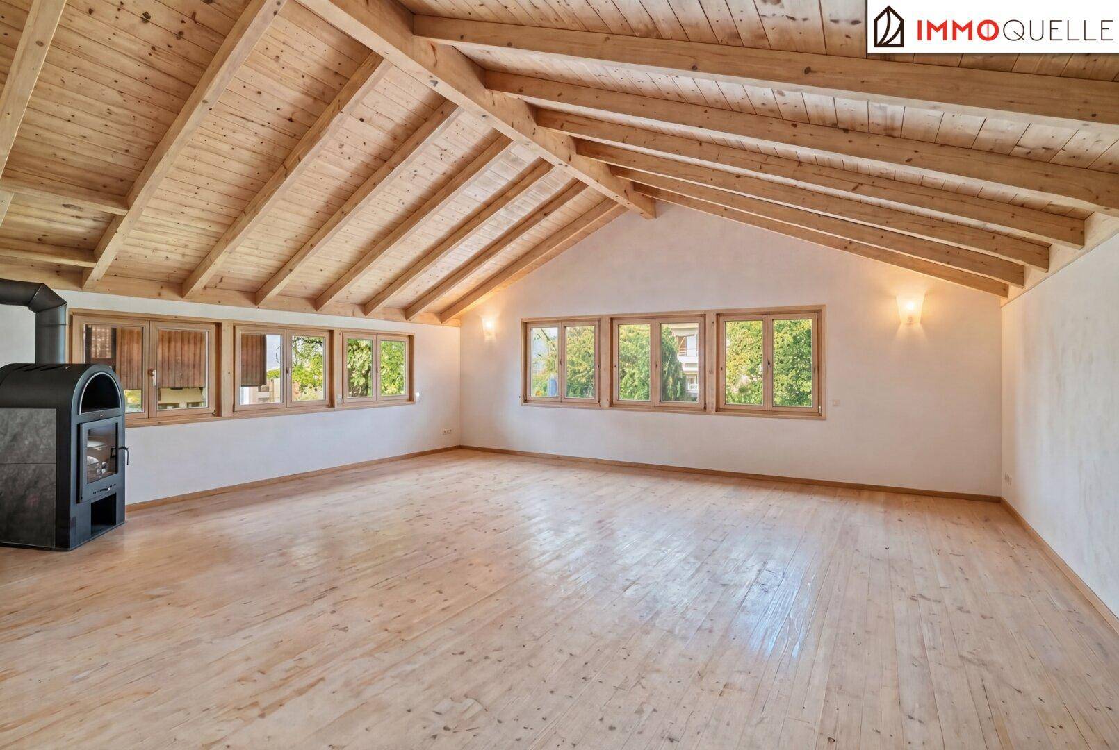 Geräumiges Dachzimmer mit Holzboden, Holzdecke, Kaminofen und vielen Fenstern.