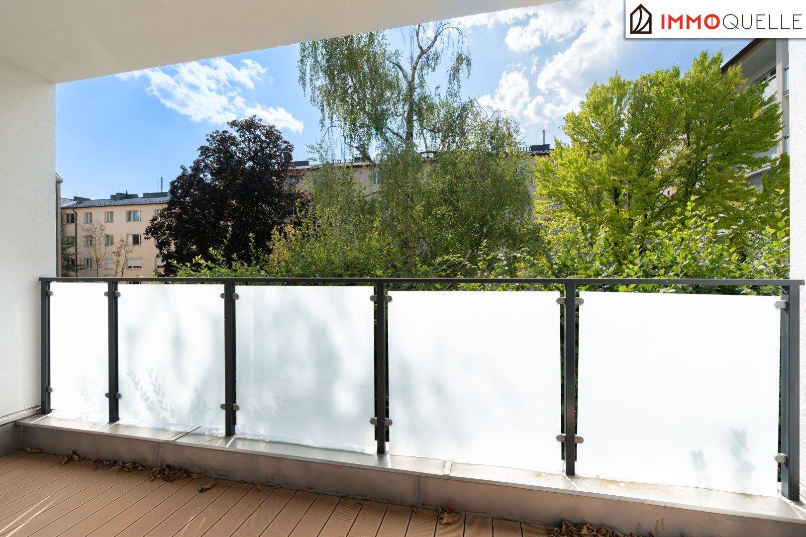 Heller Balkon mit Milchglasgeländer und Holzboden, bietet einen Blick auf Bäume und umliegende Gebäude.