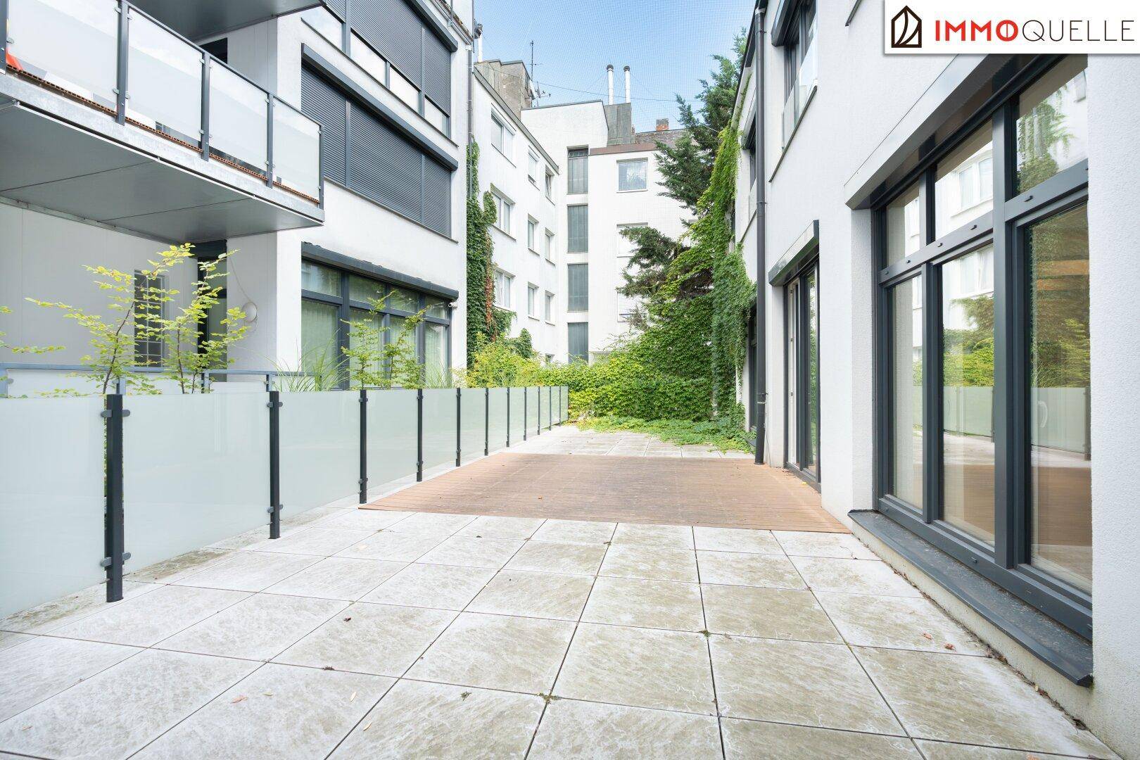 Weitläufige Terrasse im Erdgeschoss mit Steinplatten und Holzelementen, umgeben von Gebäuden und Grün.