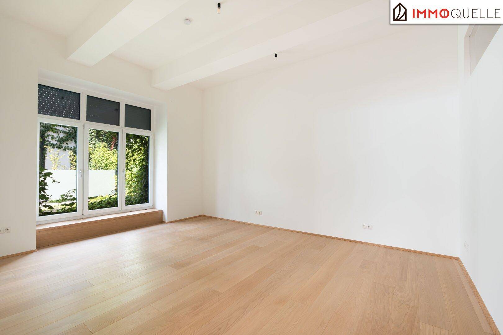 Helles, leeres Zimmer mit Holzboden und einem großen Fenster, das einen Blick ins Grüne bietet.