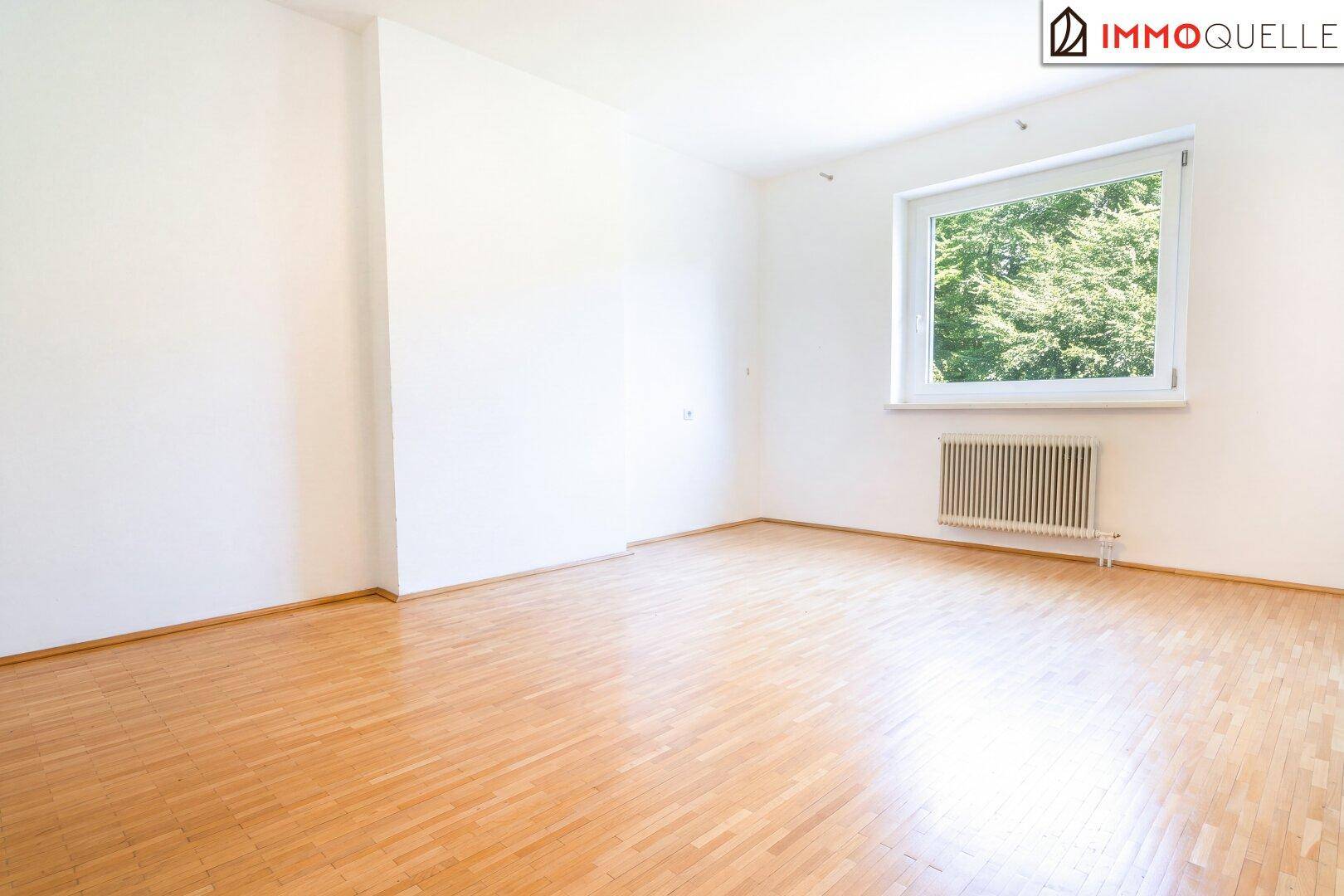 Geräumiges, leeres Schlafzimmer mit Holzboden und Fenster mit Blick ins Grüne.